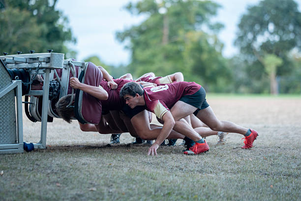 Rugby players practice together outdoors using a scrum training machine on a grass field.