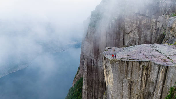 Adventurers stand atop the majestic Preikestolen Cliff, overlooking the breathtaking fjords below. Mist sweeps over the dramatic landscape, creating a captivating atmosphere.