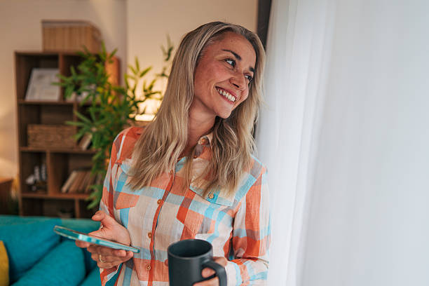 istock Smiling woman holding smartphone and coffee mug looking out window 2233554412