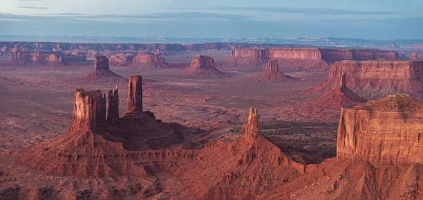 Panoramic view of Monument Valley on the Arizona–Utah border, showcasing towering sandstone buttes and mesas glowing in the soft light of sunrise across the desert landscape.