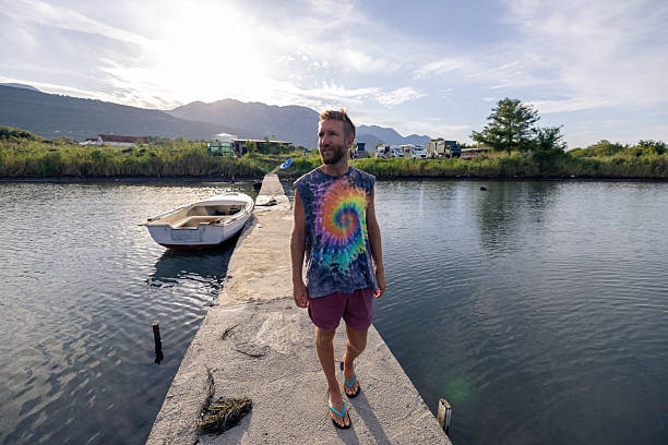 Young man in tie-dye shirt walking on narrow pier beside small boat on calm water. Casual summer scene with mountains and camper vans in background. Outdoor lifestyle and travel.