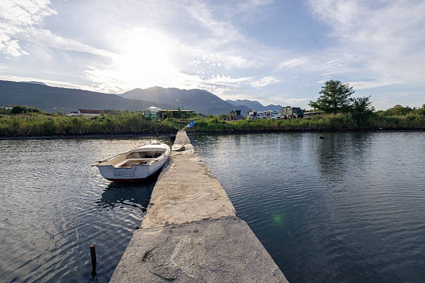 Small white boat tied to a narrow concrete pier on calm water. Rural landscape with mountains, grassy shore, and vehicles in the background. Quiet waterfront scene with natural light.