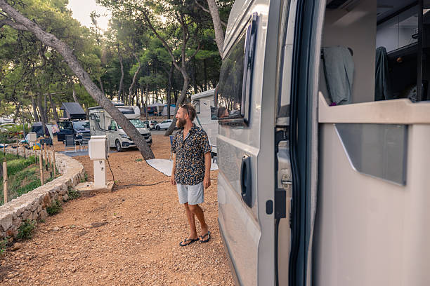 Bearded man walking on dirt path near camper van in forested campground. Casual adult enjoying outdoor camping lifestyle among parked RVs. Nature and travel adventure concept.