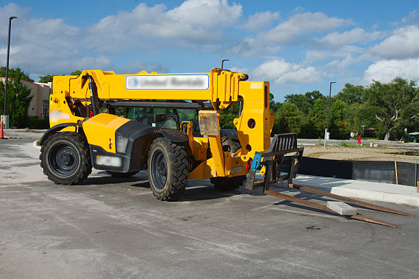Yellow construction telehandler machine with extended forks at an outdoor construction site. The vehicle illustrates industrial equipment in usage under bright daylight, surrounded by a semi-urban setting and natural greenery.