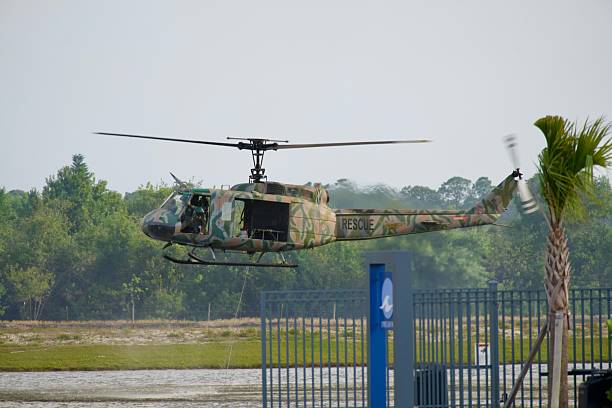 Palm Bay, FL, USA – May 16, 2025: A camouflage-painted rescue helicopter hovers low over a fenced area near a water source, preparing to assist with firefighting efforts during a large brush fire. The aircraft's crew members are visible, and the helicopter is equipped for quick deployment as the wildfire response continues in the background.