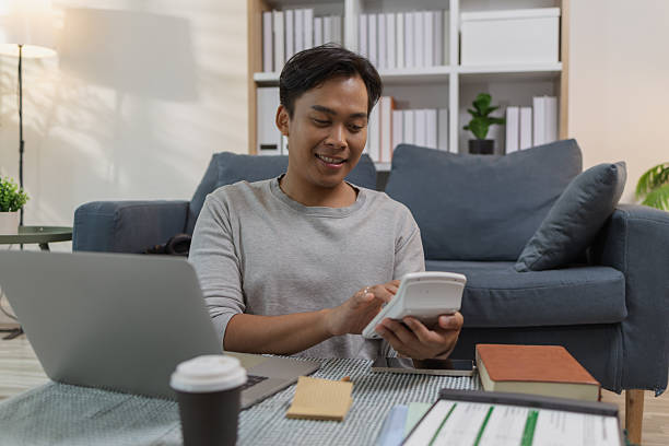 istock A young man is working at home, using a laptop and calculator with a cheerful expression. He appears relaxed and focused, enjoying his remote work in a modern living room. 2233613879
