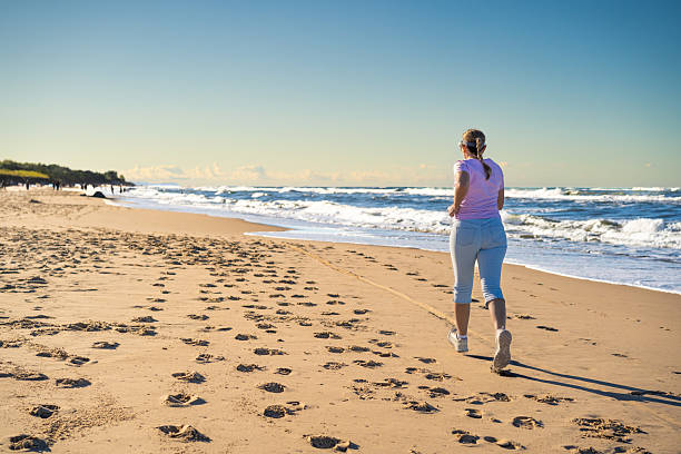 Physical activity on beach in summer. Mature woman running on sandy beach on sunny day. Back view