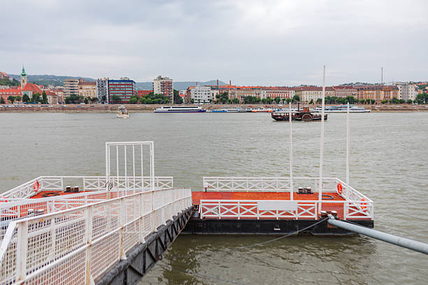 View of the Danube River in Budapest shows a docked floating pier with boats sailing across the water. In background, historic and modern buildings line the riverbank under a cloudy sky