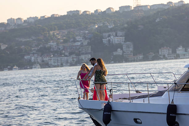 Picture of the Bosporus straight in Istanbul, Turkey, with luxury yachts cruising. A sleek motor yacht plows through sunflecked waters on Istanbul's Bosphorus Strait, leading a small flotilla toward the Sea of Marmara while bulk carriers and ferries anchor in the hazy distance. The Asian shoreline curves along the right margin, its green hillsides dotted with villas, seaside parks and whitewashed minarets, whereas silhouettes of the Historic Peninsula line the far horizon to the left. Rippling wakes, backlit spray and gentle morning glare evoke the vibrancy of summer navigation on one of the world's busiest natural waterways, underscoring Istanbul's dual identity as both leisure playground and global trade corridor linking Europe with Asia.