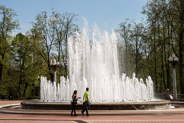 Grodno, Belarus - May 03, 2015 - Fountain in park with young adults walking on sunny day.