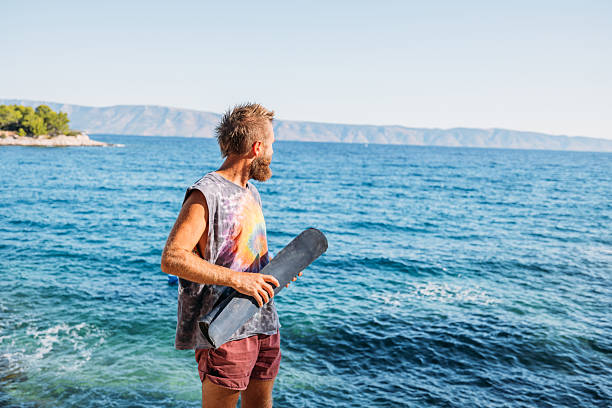 Adult man in tie-dye shirt  stands on rocky shore holding a yoga mat. Relaxed and contemplative by the ocean. Outdoor fitness and wellness lifestyle.