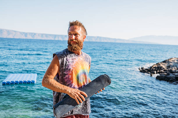 Adult man in tie-dye shirt  stands on rocky shore holding a yoga mat. Relaxed and contemplative by the ocean. Outdoor fitness and wellness lifestyle.