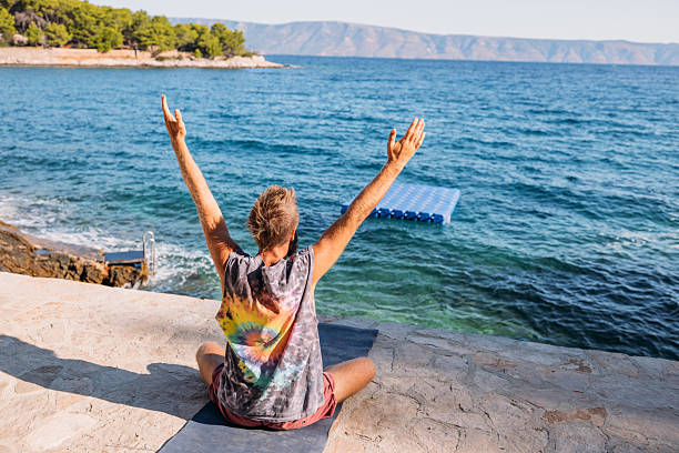 Middle-aged man sitting cross-legged on yoga mat by rocky ocean shore with arms raised. Relaxed adult practicing meditation near water. Person enjoying peaceful coastal moment.