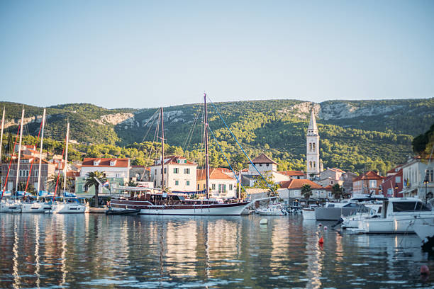 White and blue small boats moored in a calm harbor with reflections on water. Picturesque Mediterranean coastal town with historic buildings and green hills in the background. Scenic seaside village view.