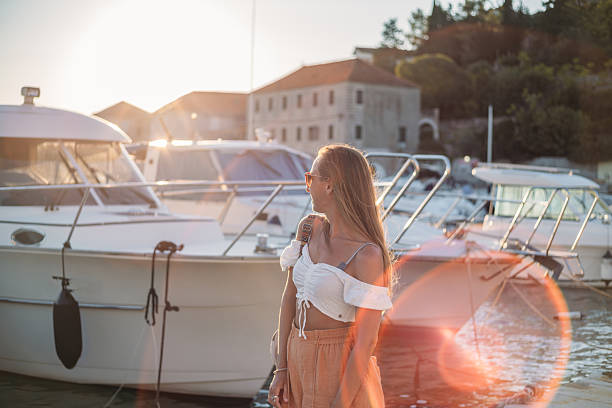 Young woman in summer outfit standing near docked white yachts at marina during sunset. Casual outdoor lifestyle by the water with boats and historic buildings in background. Leisure and travel scene.