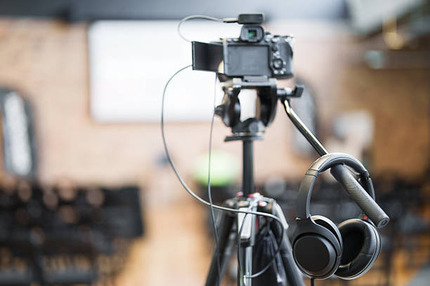 Professional filming setup with tripod stand and wireless headphones in a blurred conference hall background, capturing high-quality audio and video for events or presentations.