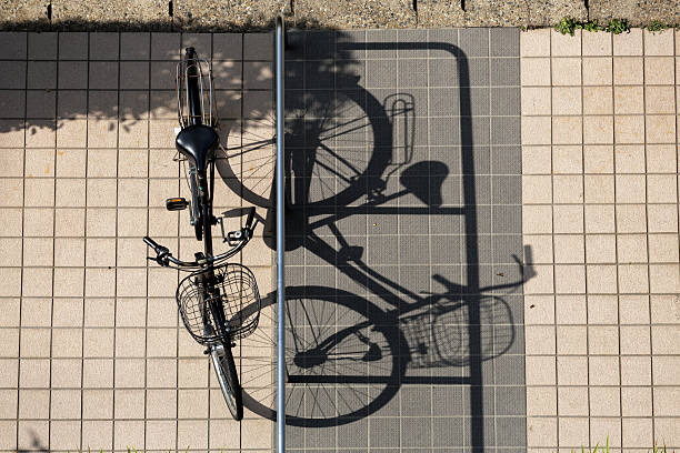 Bird-eye of black bicycle parked on tiled pavement, casting a long shadow in late afternoon sunlight. Silver railing separates area between bike and shadow, set on light beige and gray square tiles.
