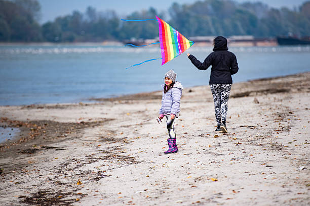 Mother and daughter embracing the chilly, windy beach day while joyfully flying a vibrant kite, creating lasting memories together