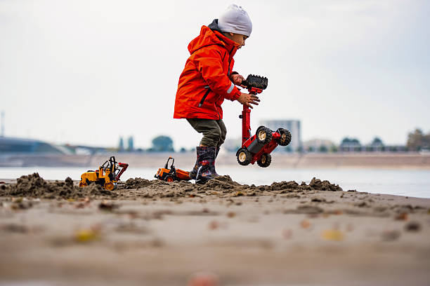Young child wearing a red jacket and rubber boots playing with toy trucks on a sandy beach near a river in autumn