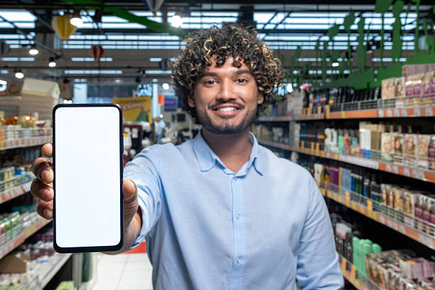 istock Man in grocery store holds up a blank smartphone screen, smiling at the camera. 2233645536