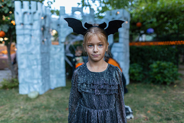 Halloween scene with a girl dressed in festive black costume and bat ears headband looking at the camera, while other children in skeleton and colorful outfits enjoy trick-or-treating in front of a cobweb-decorated castle prop outdoors.