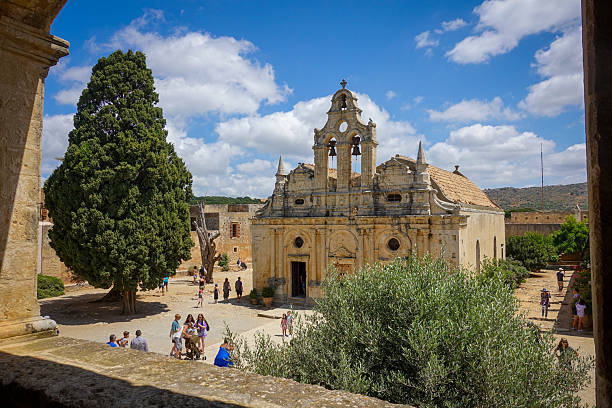 Greece, Crete, August 18, 2025: The historic Arkadi Monastery with its ornate stone facade and three-bell tower stands against a bright blue sky, surrounded by a spacious courtyard where visitors stroll near a large tree.