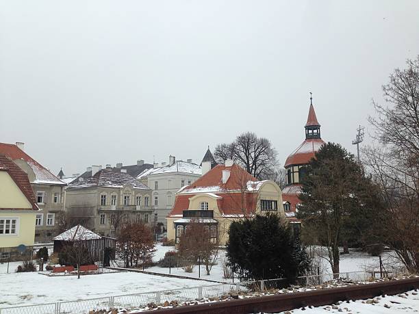 Winter scene in Krems, Austria, featuring a quaint neighborhood with snow-dusted rooftops. The buildings display traditional European architecture with multi-story houses and red-tiled roofs. A prominent church tower with a pointed, pyramid-shaped top stands amidst bare trees. The ground is lightly covered with snow, and a garden area is visible with bare bushes and trees. The sky is overcast, creating a tranquil yet chilly atmosphere.