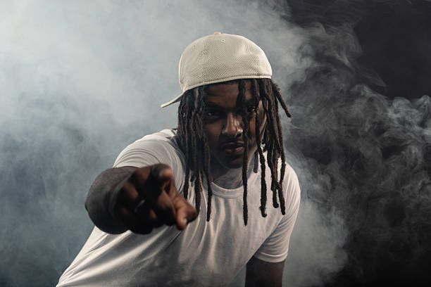 man with dreadlocks wearing a hat and white shirt posing for the camera in a studio against a smoky background. Confident and serious young man.