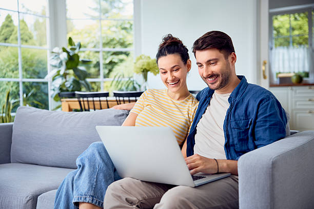 istock Young couple sitting on sofa at home, smiling while using laptop in living room 2233659968