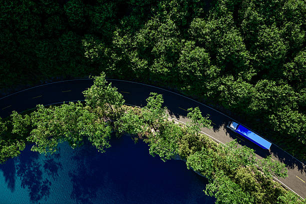 Aerial image of a blue cargo truck on a forest road near a body of water, surrounded by lush green forest. The vivid blue and green contrast, along with the clean lines of the road and bridge, emphasizes themes of transportation, infrastructure, and eco-conscious logistics.