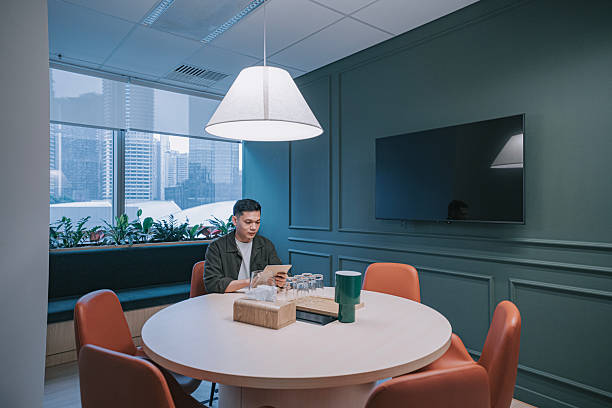 Asian young man sitting alone at a round table in a modern office meeting room, using a digital tablet with city buildings visible through the window