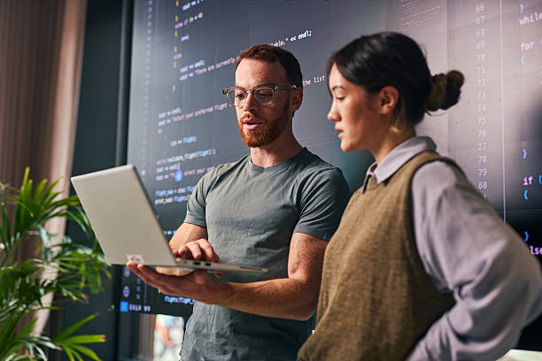 two young adult programmers discussing code on laptop in office - programador imagens e fotografias de stock