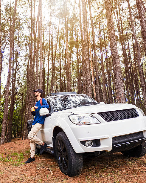 A stylish young man enjoy a travel break with their SUV in a scenic pine forest. The scene creating a moment of connection and lifestyle inspiration. The bright daylight and natural environment highlight themes of freedom, adventure, and outdoor travel. Perfect image for concepts of road trips, camping, tourism, and active lifestyle.