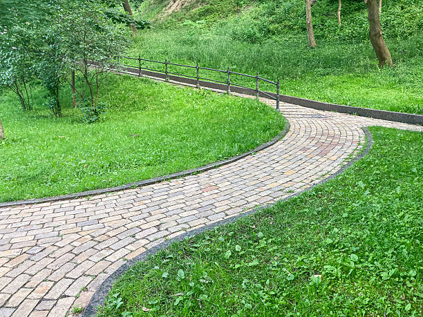 istock Curving brick path through a park with neat green bushes, leading into a lush forest under overcast daylight t 2233683286