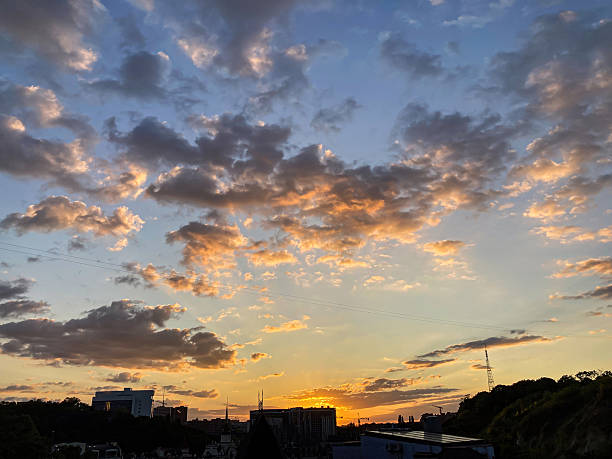 Tranquil urban sunset, dense fluffy clouds, orange and pink hues, textured sky backdrop, silhouetted buildings, reflective body of water, soft diffused lighting, long shadows, low angle view