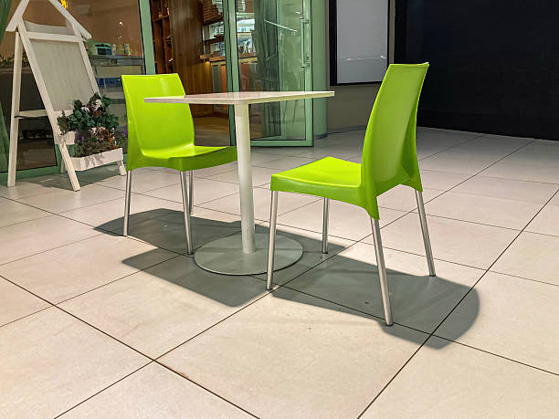 Modern indoor setting possibly cafe or restaurant, two vibrant yellow chairs, round white table, contemporary design, metallic legs, natural lighting, rectangular tiles, shelvesstorage units back