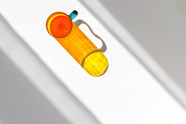 Top view of yellow orange colored glass mug creating vivid light reflections and caustics on white background. Transparent cup at sunlight with window shadow, aesthetic summer drink concept, copyspace