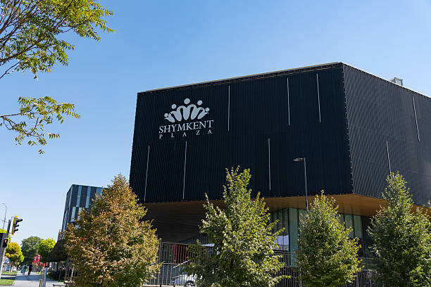 Shymkent, Kazakhstan, August 30, 2025: Contemporary black facade of Shymkent Plaza shopping mall with crown logo, urban street view, green trees, and clear blue sky