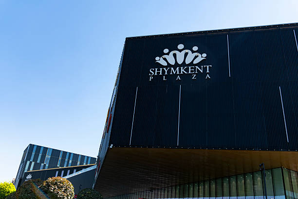 Shymkent, Kazakhstan, August 30, 2025: Contemporary black facade of Shymkent Plaza shopping mall with crown logo, urban street view, green trees, and clear blue sky