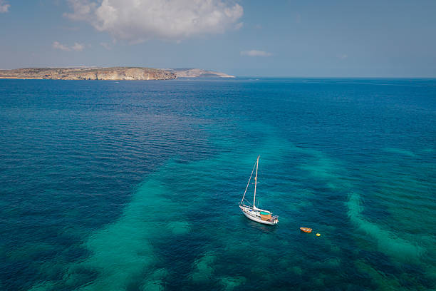 A white sailboat floats peacefully in the turquoise waters of the open sea with a small dinghy nearby. The distant rocky coastline and blue sky create a tranquil and scenic seascape.