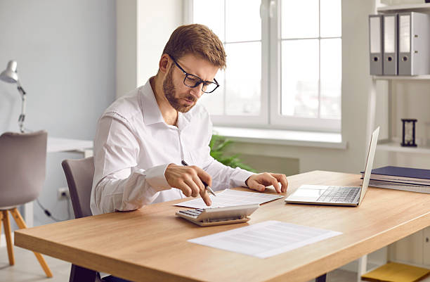 istock Young focused serious business man working on laptop at office making calculations. 2233695497
