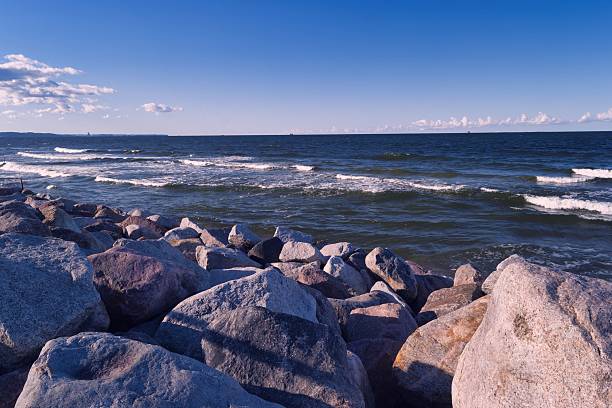 The coastline features large, smooth rocks scattered along the shore. Waves gently roll in under a bright blue sky with scattered clouds, capturing the essence of a serene seaside atmosphere.