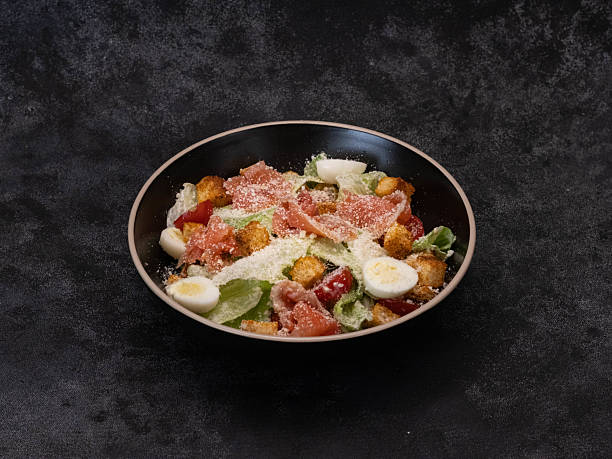 Enjoy a colorful Caesar salad with salmon,lettuce leaves, quail eggs, fresh veggies, cherry, and crispy toppings for added texture and flavor on black background.