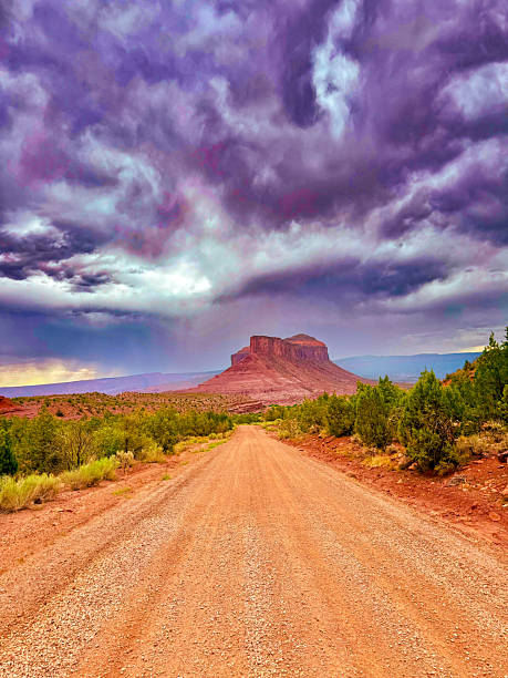 A dramatic desert road leads the eye directly to a solitary red rock mesa rising from the arid terrain. Above, an intensely colored sky swirls with violet and steel-gray storm clouds, suggesting an impending downpour. The red gravel road is flanked by hardy desert shrubs and junipers, adding depth and contrast to the rugged scene. This photo captures the raw and cinematic beauty of the American Southwest, where weather and geology collide in powerful visual harmony.