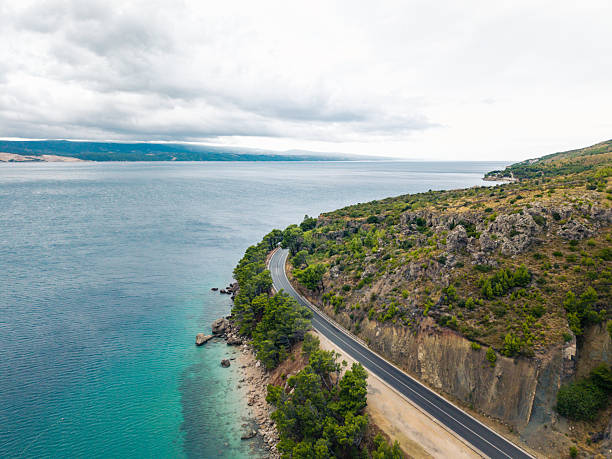A winding coastal road runs beside clear turquoise water and a rocky hillside covered with green trees. Scenic landscape with beach, ocean, and natural vegetation along the shore. Coastal highway view.