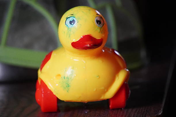 Yellow rubber duck toy sitting on a white balcony wall, facing forward with a blurred bright blue sky, the sea, and green trees in the background, creating a relaxed summer holiday mood