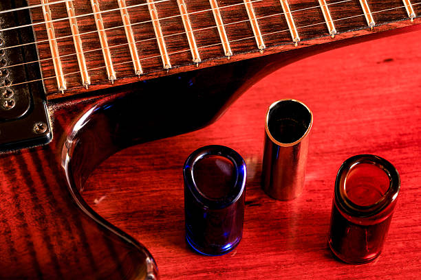 Three electric guitar slides, two made of glass and one of metal are placed by the side of an electric guitar on a shiny rosewood table. No people. Tight composition.
