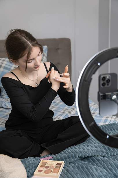istock Teenager influencer sitting on bed, applying makeup, and filming tutorial video using smartphone with ring light and sharing tips with followers 2233723964