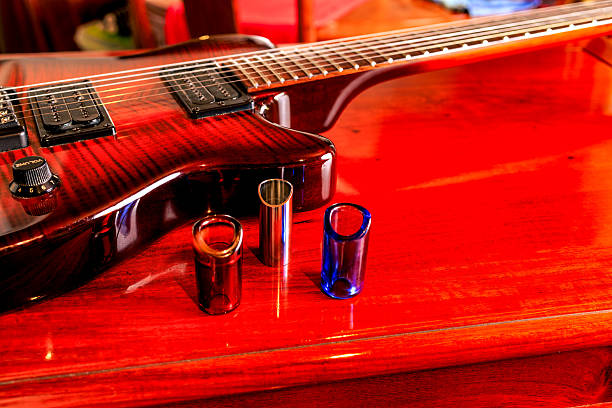 A headless electric guitar, evident by the special bridge, is placed on a shiny rosewood table. Three guitar slides are seen to the centre of the image. Two are made of glass and one of metal. The high degree of polish on the table produces reflection. Studio shot with copy space.