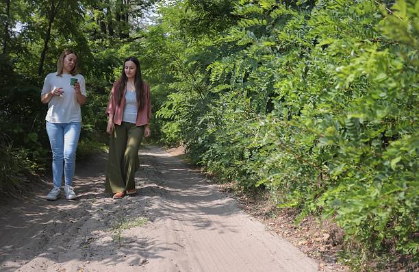 Two women walk along a sandy forest path surrounded by green trees, one holding a cup. They enjoy a calm summer day together in nature.